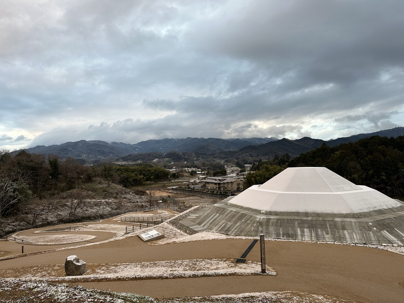 薄く雪の積もった牽牛子塚古墳。復元された八角形の墳丘と、奥に広がる明日香の集落と山並み
