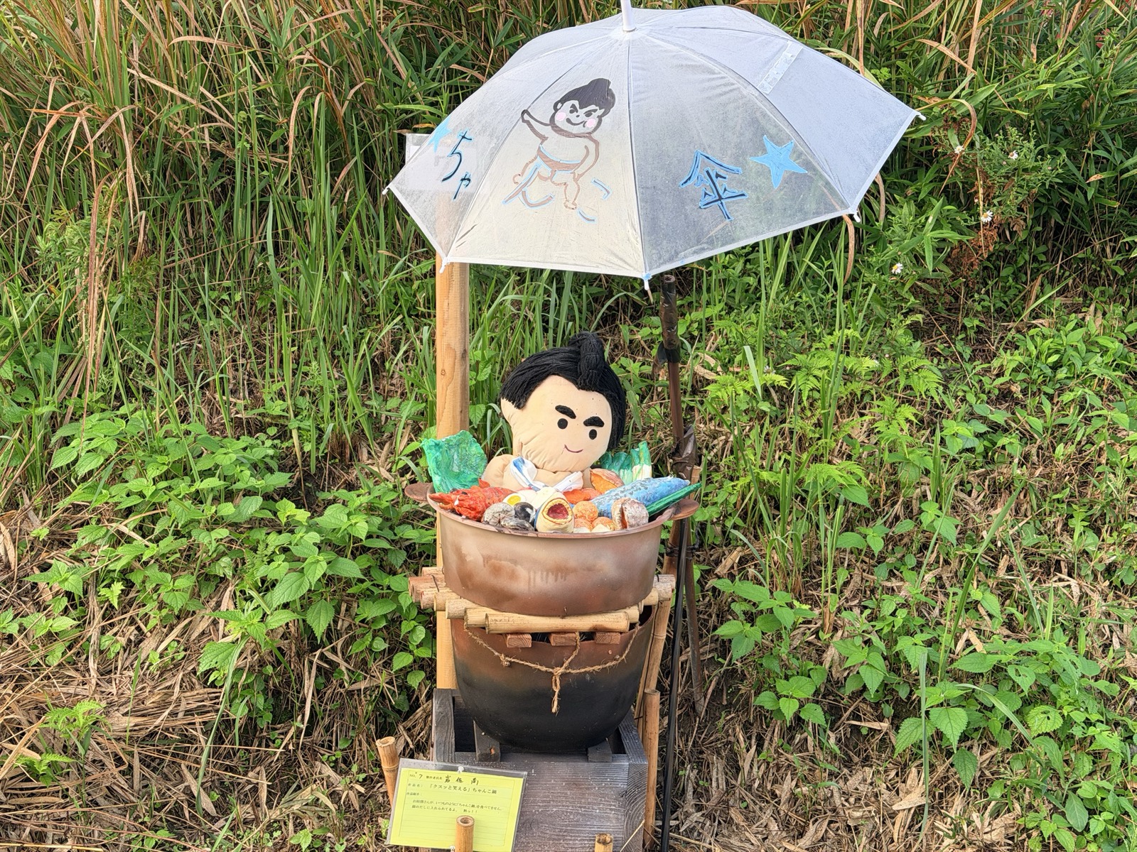 鍋のなかに浸かった力士の案山子。頭上に手描きの傘がさしかけられている