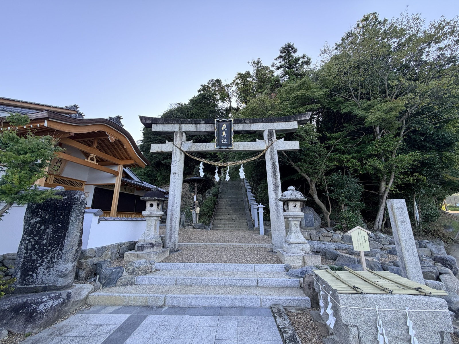 The shrine entrance, with stone steps rising beyond a stone torii, the gathering hall on the left and the Asuka Well on the right