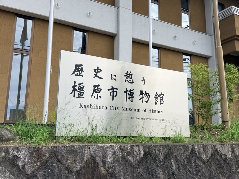 「歴史に憩う橿原市博物館 Kashihara City Museum of History」と刻まれた館名板と、奥に建つ博物館の建物