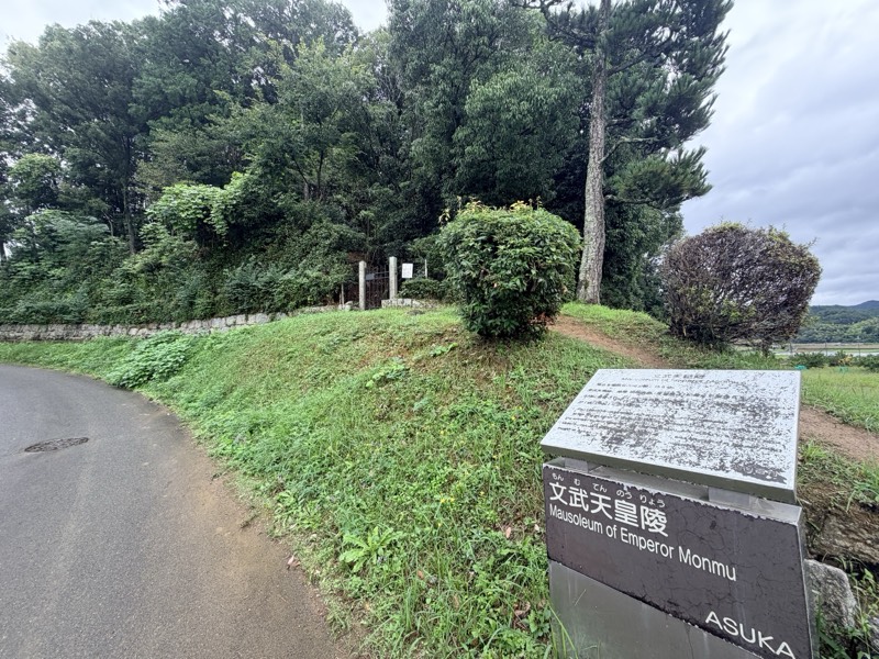 道路沿いの墳丘と「文武天皇陵」の説明板。石垣の上に拝所の門が見える