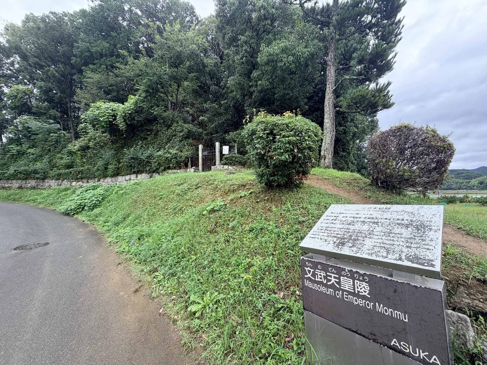 道路沿いの墳丘と「文武天皇陵」の説明板。石垣の上に拝所の門が見える