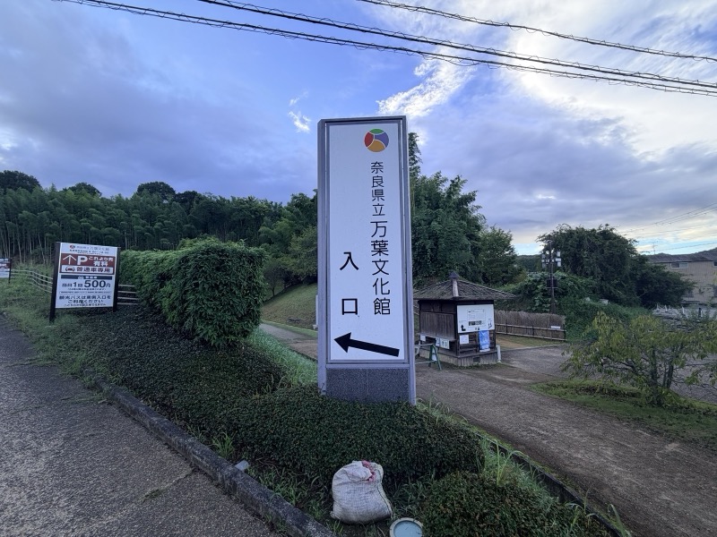 「奈良県立万葉文化館 入口」と書かれた案内塔。奥に竹林と受付の小屋