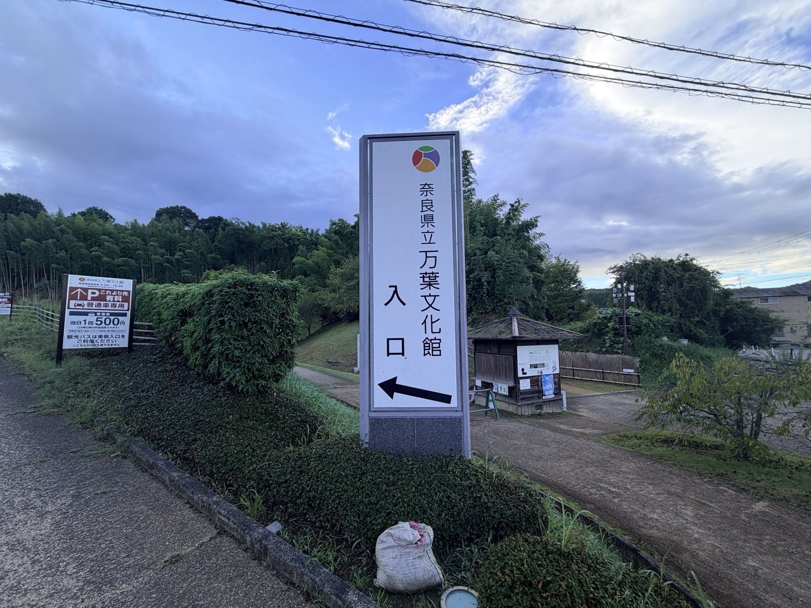 「奈良県立万葉文化館 入口」と書かれた案内塔。奥に竹林と受付の小屋