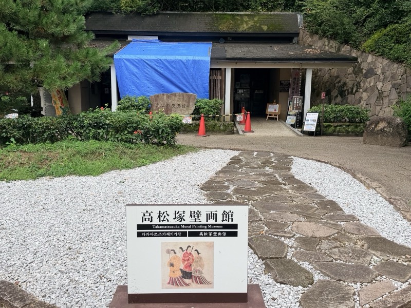 高松塚壁画館の外観。白い玉砂利の前に、女子群像の絵を添えた館名の看板が立つ